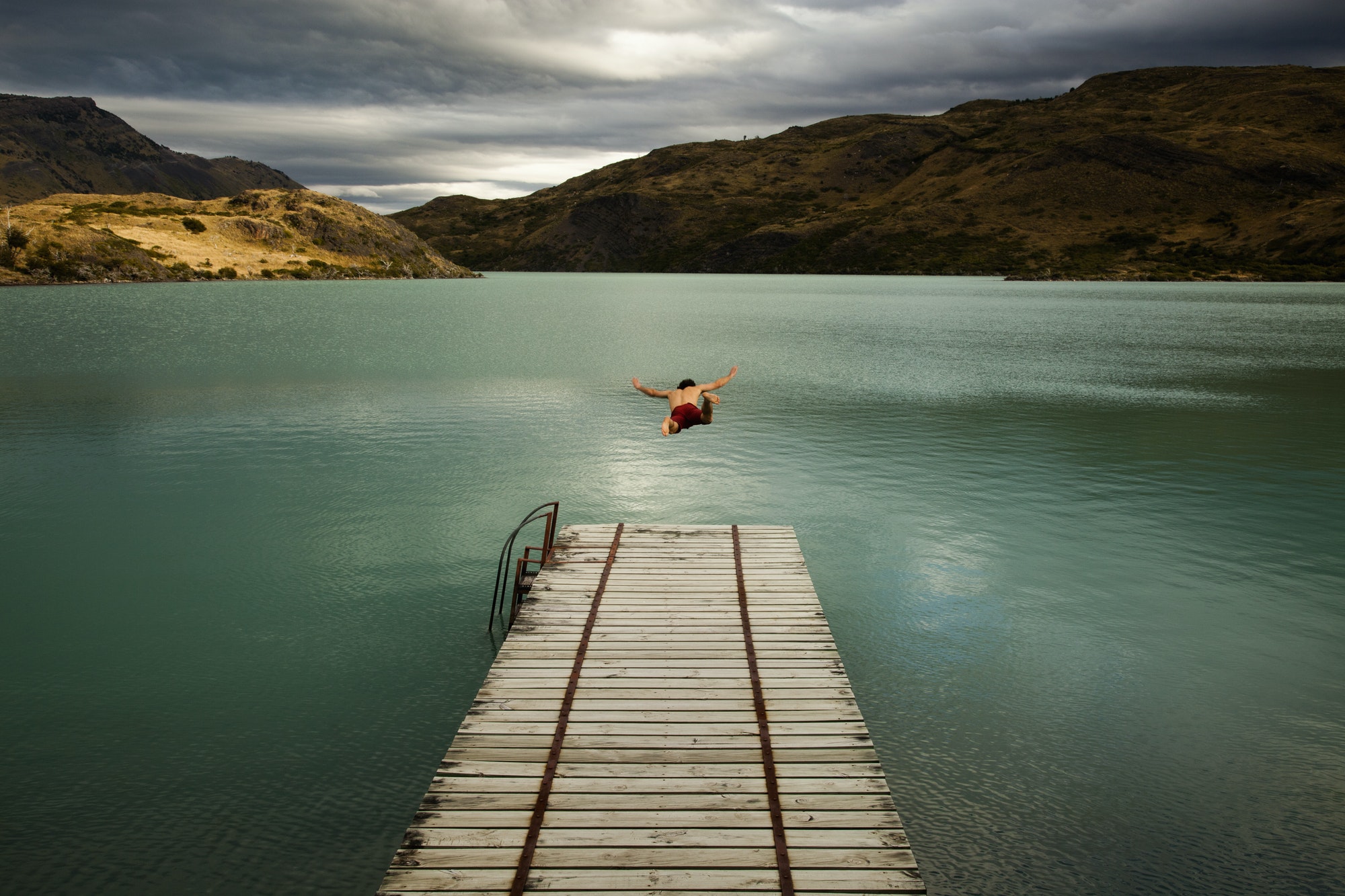A young man in mid air, diving off a wooden pier, into calm lake, Chile.